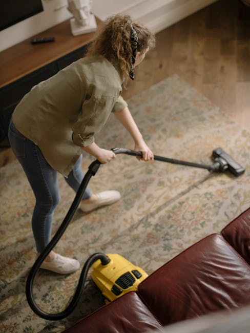 A woman with curly hair, wearing a beige shirt and dark jeans, is using a yellow handheld vacuum cleaner to deep clean a patterned area rug in a living room. The room features wooden flooring partiall
