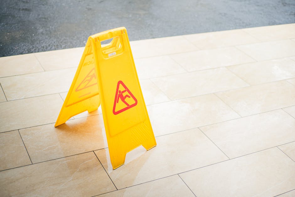 A yellow 'wet floor' caution sign is placed on a smooth, beige tiled floor in an indoor setting, indicating temporary surface cleaning or mopping. The tiles are clean and shiny, with natural light hig
