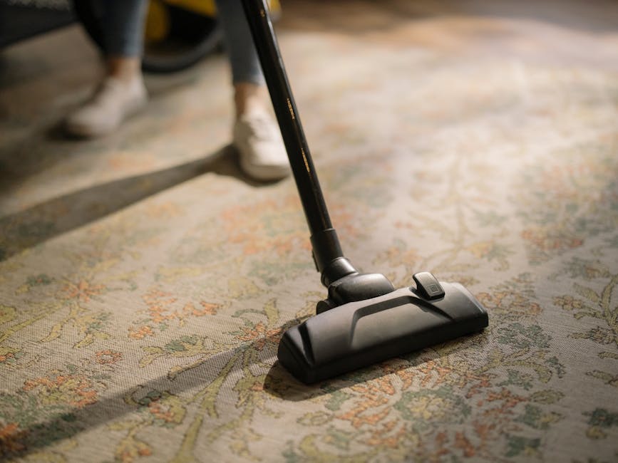 A close-up view of a vacuum cleaner head actively cleaning an ornate, patterned area rug with floral and vine motifs in muted colors, on a residential or commercial floor. The vacuum attachment is bla